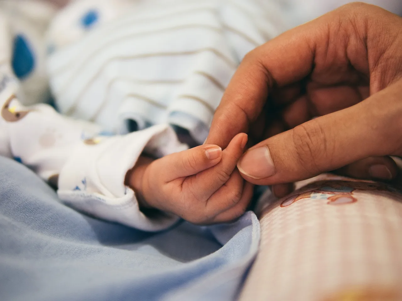Közelről egy csecsemő apró keze látható, amint egy felnőtt ujját megfogja; a baba csíkos ruhában, kék takarón fekszik.