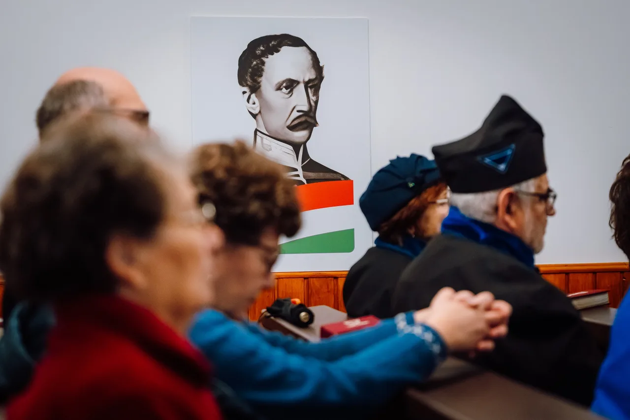 Idősebb emberek ülnek egy templom padsoraiban, háttérben egy férfi portréját ábrázoló plakát látható piros-fehér-zöld sávval, mellette egy fekete sapkás férfi és társai figyelnek.