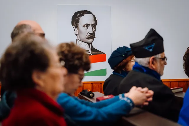 Idősebb emberek ülnek egy templom padsoraiban, háttérben egy férfi portréját ábrázoló plakát látható piros-fehér-zöld sávval, mellette egy fekete sapkás férfi és társai figyelnek.