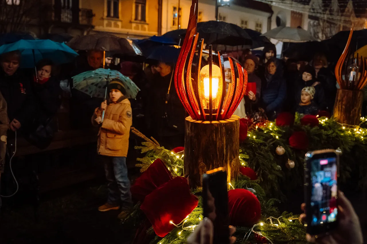 Esti adventi gyertyagyújtás fenyőgirlanddal és piros bársonydíszekkel díszített téren; egy láng formájú, fából és üvegből készült lámpa világít középen, körülötte esernyőt tartó emberek állnak esőben, előtérben telefonnal fotózó kéz látszik.
