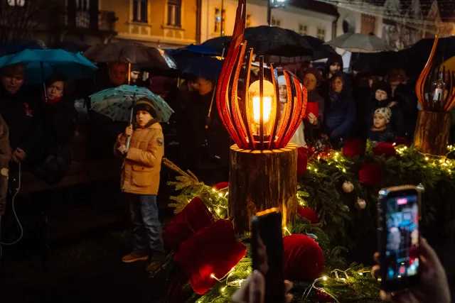 Esti adventi gyertyagyújtás fenyőgirlanddal és piros bársonydíszekkel díszített téren; egy láng formájú, fából és üvegből készült lámpa világít középen, körülötte esernyőt tartó emberek állnak esőben, előtérben telefonnal fotózó kéz látszik.