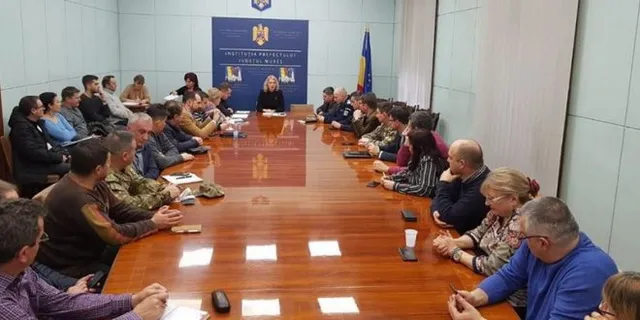 Hosszú tárgyalóasztal körül ülő emberek egy tanácskozáson, hátul a Maros Megyei Prefektúra emblémás kék háttere és Románia zászlaja látható, katonai és rendőri egyenruhás résztvevőkkel.