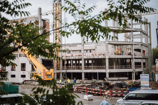 Építkezés látható lombos ágakon átfotózva: betonvázas, több emeletes épület szerkezete, sárga mobildaru és toronydaru, munkások dolgoznak a szinteken és a földszinten, előtérben munkagépek, vasanyagok, korlátok és egy CONA feliratú tábla, a háttérben lakótömbök.