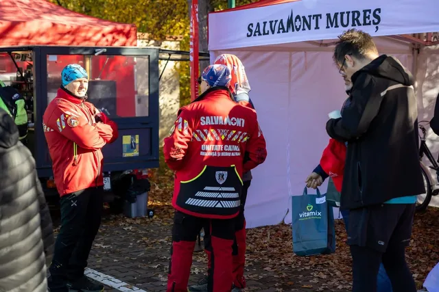 Piros Salvamont Mureș egyenruhás hegyimentők állnak egy sátras standnál, beszélgetnek egymással és két másik emberrel, a háttérben egy fehér sátor Salvamont Mureș felirattal és egy sötét szervizkocsi látható.