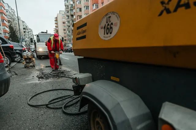 Utcán piros overallos, sárga láthatósági mellényt viselő munkás légkalapáccsal bontja az aszfaltot, mellette sárga Atlas Copco kompresszor áll, háttérben tömbházak és egy teherautó.