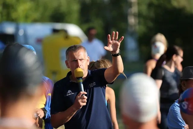 Egy férfi sárga mikrofonnal a kezében beszél és felemelt kézzel int, körülötte sportolók és nézők állnak egy szabadtéri rendezvényen.