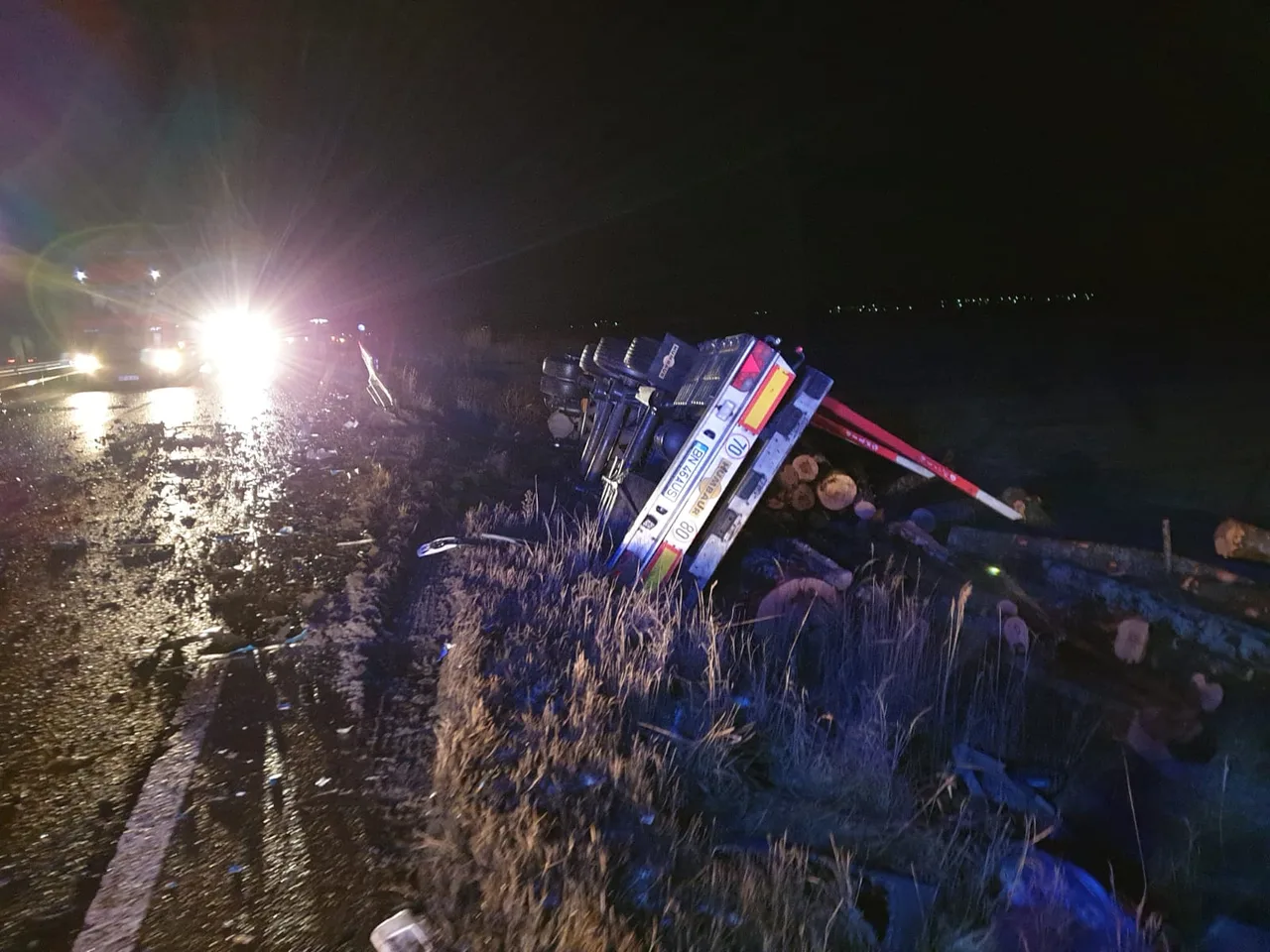 Éjszakai közúti balesetjelenet: egy fát szállító nyergesvontató az árokban felborulva hever, rakománya (fatörzsek) szétszóródott mellette. Háttérben a mentőalakulatok fényszórói világítják meg az esőáztatta útszakaszt.