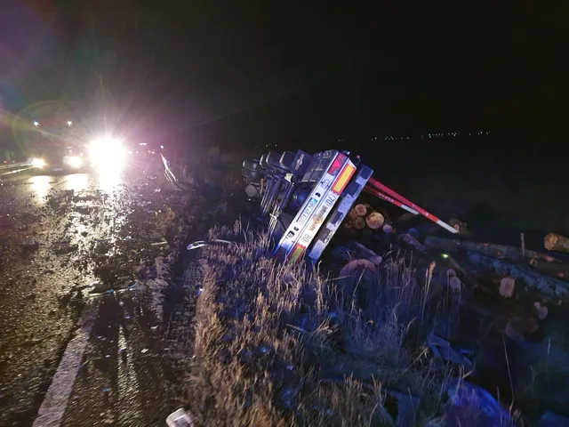 Éjszakai közúti balesetjelenet: egy fát szállító nyergesvontató az árokban felborulva hever, rakománya (fatörzsek) szétszóródott mellette. Háttérben a mentőalakulatok fényszórói világítják meg az esőáztatta útszakaszt.