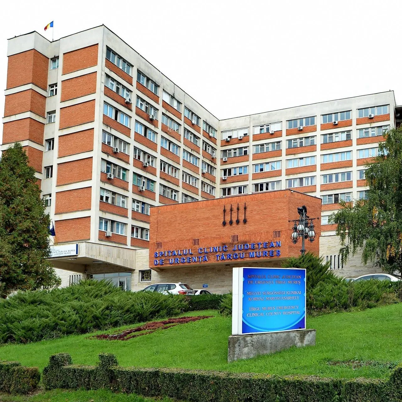 A Maros Megyei Sürgősségi Klinikai Kórház (Spitalul Clinic Județean de Urgență Târgu Mureș) modern, tégla- és fehér burkolatú épülete látható, előtte kék háromnyelvű felirati tábla, nyírt sövény és zöld gyep, néhány parkoló autóval.