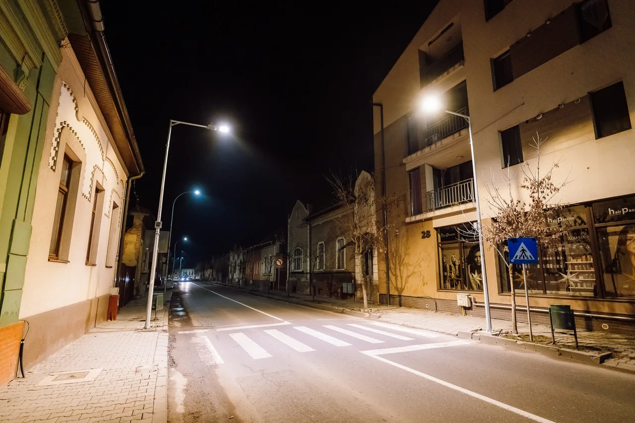 Éjszakai utcakép Marosvásárhelyen, üres gyalogátkelővel, amelyet erős lámpafény világít meg. A háttérben régi és újabb épületek, üzletek láthatók, az utcát utcai lámpák világítják meg.