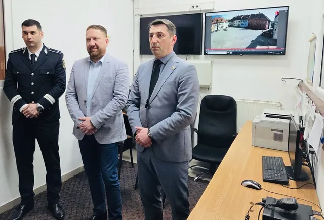 Három férfi áll egy irodahelyiségben, balról egy egyenruhás rendőr, középen és jobbra öltönyös férfiak, mögöttük fali monitorok láthatók, egyiken egy térfigyelő kamera felvétele egy régi városközpontról.