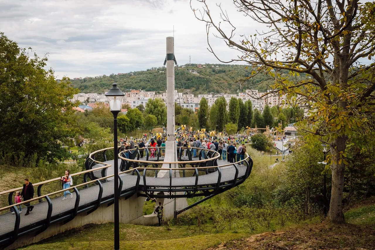 Kilátás egy magasan vezetett hídszerű gyalogos sétányra a Cserealja parkban, amelyen sokan sétálnak; a háttérben lakótömbök, erdős domboldal és játszótér látható.