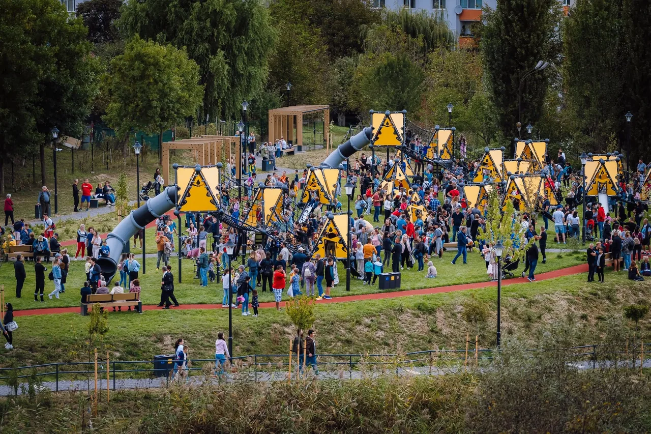 Madártávlati kép egy modern kalandparkos játszótérről, ahol nagy sárga háromszög alakú táblák és kötélmászó elemek között sok ember, köztük gyerekek és felnőttek sétálnak és játszanak. A háttérben fák, egy lakóház és favázas pergolák láthatók.
