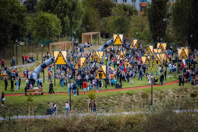 Madártávlati kép egy modern kalandparkos játszótérről, ahol nagy sárga háromszög alakú táblák és kötélmászó elemek között sok ember, köztük gyerekek és felnőttek sétálnak és játszanak. A háttérben fák, egy lakóház és favázas pergolák láthatók.