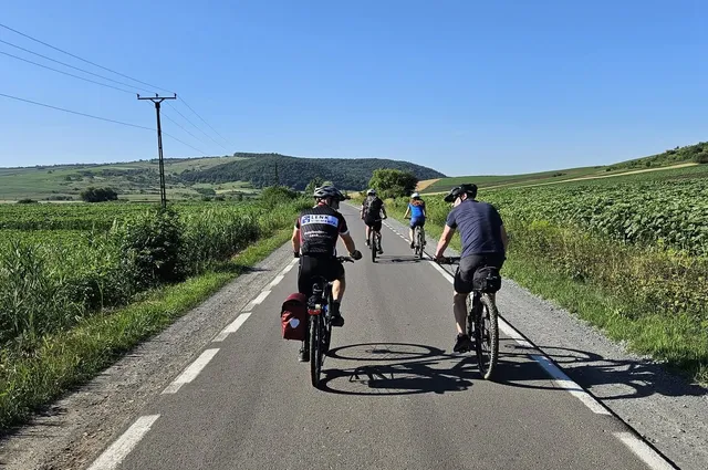 Négy kerékpáros halad egy vidéki aszfaltúton, mezők és dombok között, napos időben.