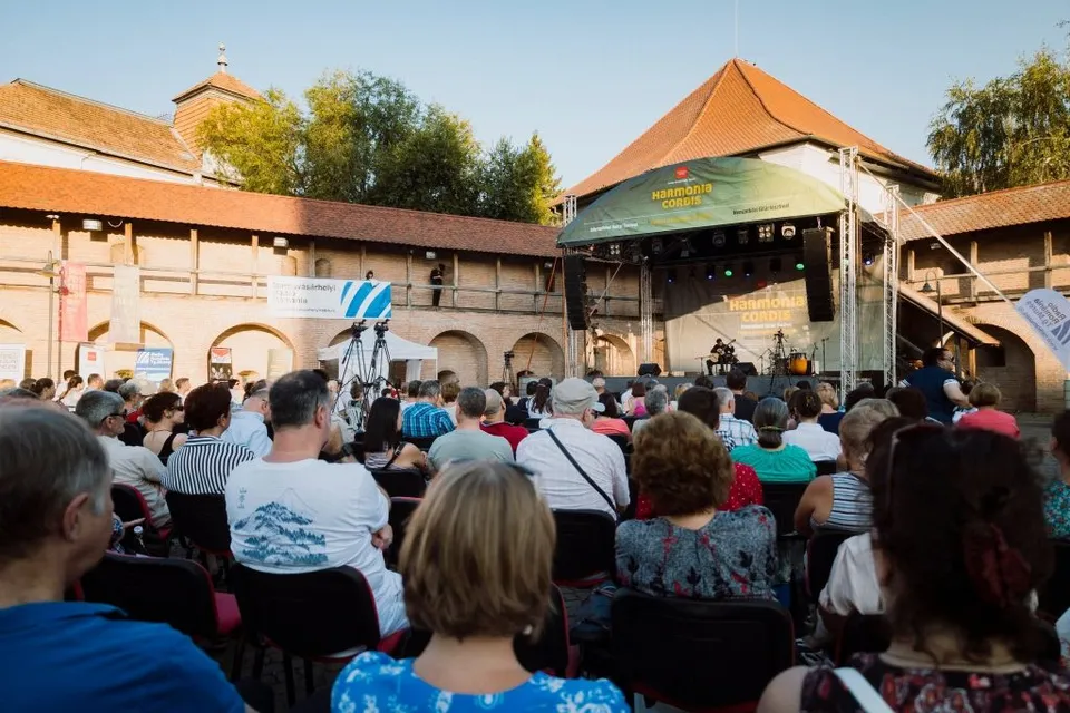 Szabadtéri koncert a marosvásárhelyi vár udvarán, ülő nézőközönséggel, háttérben színpaddal, ahol zenészek játszanak, és a vár bástyafalai láthatók.