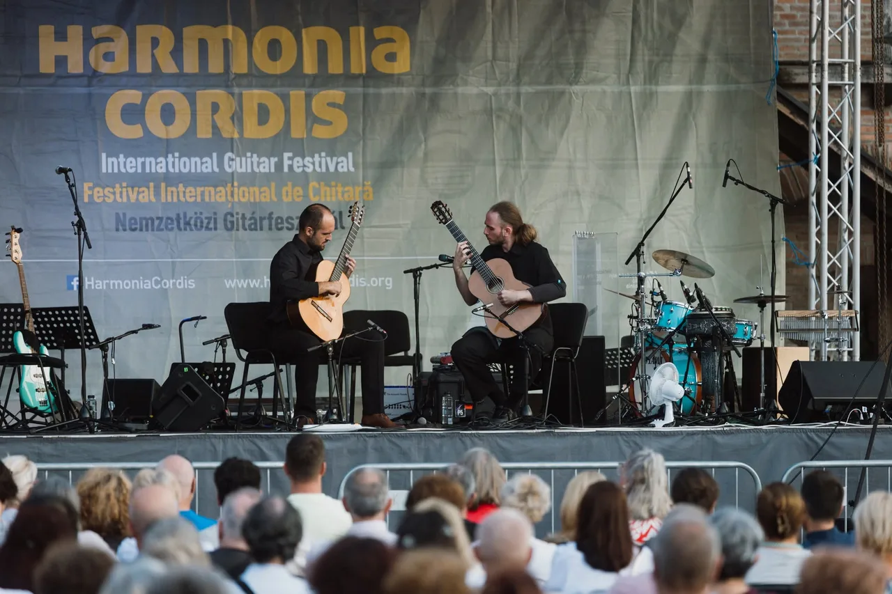 Két gitáros ül egymással szemben a színpadon, feketébe öltözve, klasszikus gitáron játszanak a Harmonia Cordis Nemzetközi Gitárfesztivál koncertjén; háttérben a fesztivál felirata, dobfelszerelés és hangtechnikai eszközök, előtérben ülő közönség figyeli az előadást.