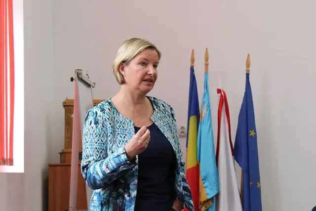 Egy szőke nőt, mintás kardigánban és sötét ruhában, beszéd közben, kezével gesztikulálva egy teremben, mögötte román, székely, magyar és európai uniós zászlók.