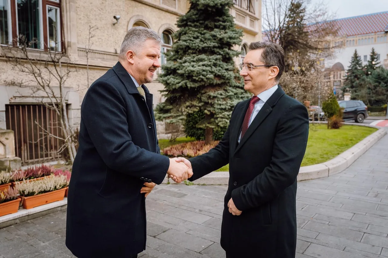 Két öltönyös, sötét kabátot viselő férfi kézfogással köszönti egymást egy épület előtti sétányon, háttérben fenyőfák, egy régi stílusú épület és zöld gyep látható.