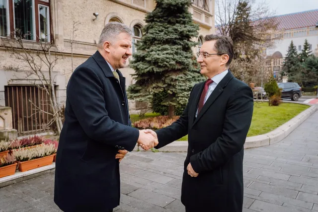 Két öltönyös, sötét kabátot viselő férfi kézfogással köszönti egymást egy épület előtti sétányon, háttérben fenyőfák, egy régi stílusú épület és zöld gyep látható.