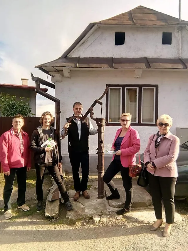Öt ember áll egy régi kézi vízpumpa körül egy ház előtt, ketten piros/rózsaszín kabátban, egy férfi a pumpát fogja, mögöttük fehér falú, cserepes tetős épület.