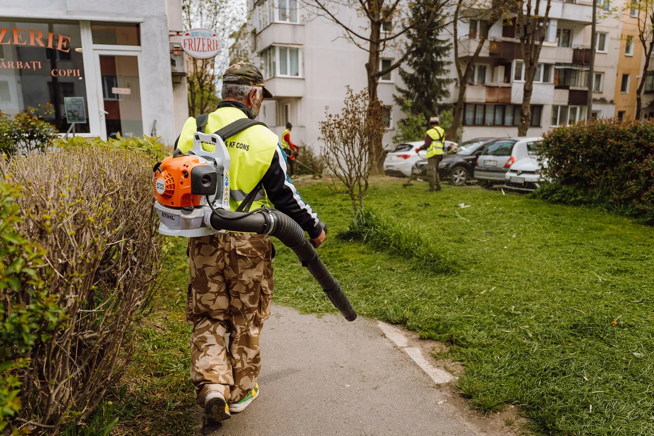 Fényvisszaverő mellényt viselő férfi hátizsákos lombfúvóval sétál egy tömbház sétányán, a háttérben zöld gyepen egy másik munkás fűkaszával dolgozik, parkoló autók és lakóházak láthatók.