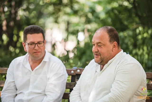 Két férfi ül egy fapadon, kültéren, zöld lombos háttér előtt; mindketten fehér inget viselnek, a jobb oldali férfi beszél, a bal oldali szemüveges férfi figyelmesen hallgatja.