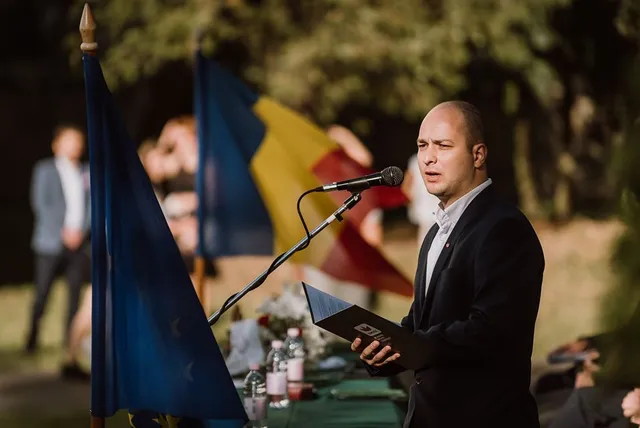 Öltönyös férfi beszél egy kültéri rendezvényen mikrofonba, kezében jegyzetfüzetet tart, mellette EU-s és román zászlók, háttérben zöld terítős asztal és fák.
