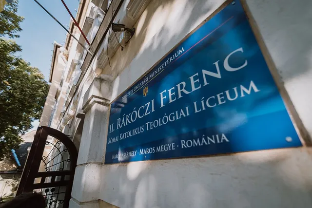 Kék háttérszínű intézményi tábla látható egy épület falán, rajta 'II. Rákóczi Ferenc Római Katolikus Teológiai Líceum, Marosvásárhely · Maros megye · Románia' felirat, mellette egy vaskovácsolt kapu és fa.