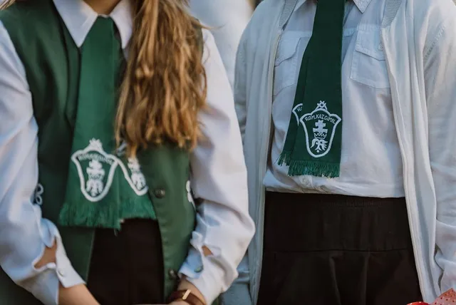 Két fiatal áll egymás mellett, mindketten fehér inget és sötétzöld, rojtos végű, címeres kendőt (sálat) viselnek nyakukban, az egyikük sötétzöld mellényt is hord.