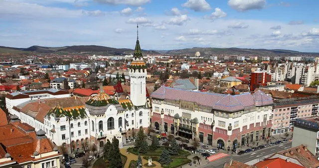 Légi felvétel Marosvásárhely belvárosáról, középen a Kultúrpalota tornyával és a jellegzetes mázas cseréptetős Városháza-épülettel, körülöttük a régi városrész tetői és háttérben dombok láthatók.