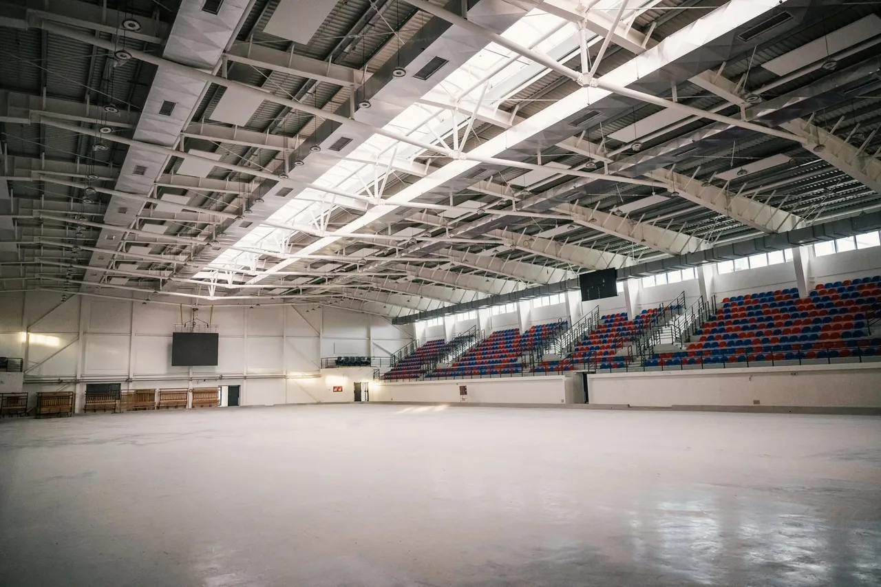 Nagy, üres beltéri sportcsarnok jégpálya nélküli, sima betonpadlóval, jobb oldalon kék-piros lelátói székekkel, felül fémszerkezetes tetővel és tetőablakokkal.