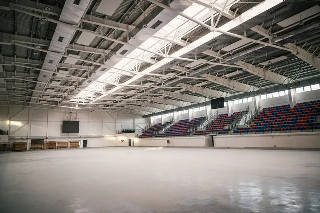 Nagy, üres beltéri sportcsarnok jégpálya nélküli, sima betonpadlóval, jobb oldalon kék-piros lelátói székekkel, felül fémszerkezetes tetővel és tetőablakokkal.
