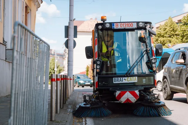 Utcai kefés seprőgép áll egy járda mellett, forgó kefékkel elöl, sofőrje sárga láthatósági mellényben ül a vezetőülésben; az oldalán „Brai-Cata” felirat, mögötte parkoló autók és egy fém korlát.