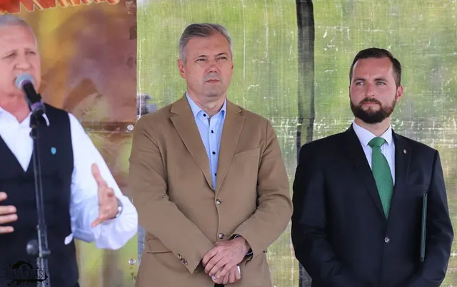 Három férfi áll egy szabadtéri rendezvényen, közülük az egyik mikrofonba beszél és gesztikulál, a másik kettő mellette áll, hallgatja.