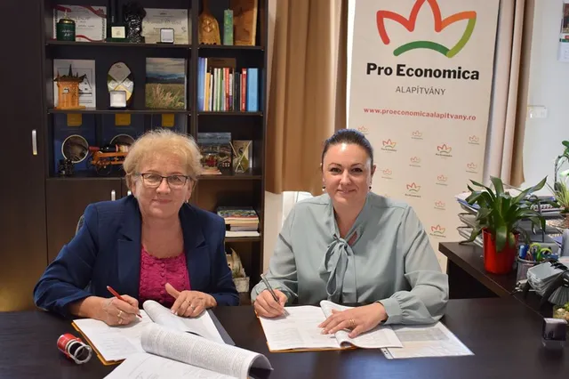 Két nő ül egy íróasztalnál irodában, mindketten dokumentumokat írnak alá, mögöttük egy 'Pro Economica Alapítvány' feliratú roll-up banner látható.