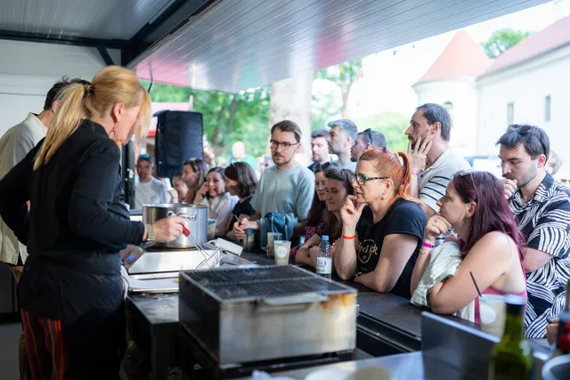 Fedett standnál egy szőke hajú nő főzőbemutatót tart egy fém edény és grillrácsok mellett, körülötte sok néző figyeli szorosan a bemutatót egy fesztiválon.