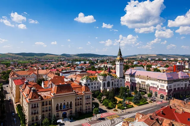 Marosvásárhely belvárosának légi felvétele napsütésben, látható a Kultúrpalota tornya, a Rózsák tere színes mozaikcsempés épülettel, és a városközpont jellegzetes tetőzete zöld dombok háttere előtt.
