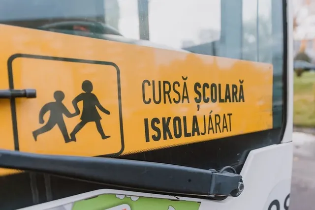 Egy busz szélvédőjén sárga tábla látható, rajta gyerekeket ábrázoló piktogram és román-magyar felirat: „Cursă școlară / Iskolajárat”.