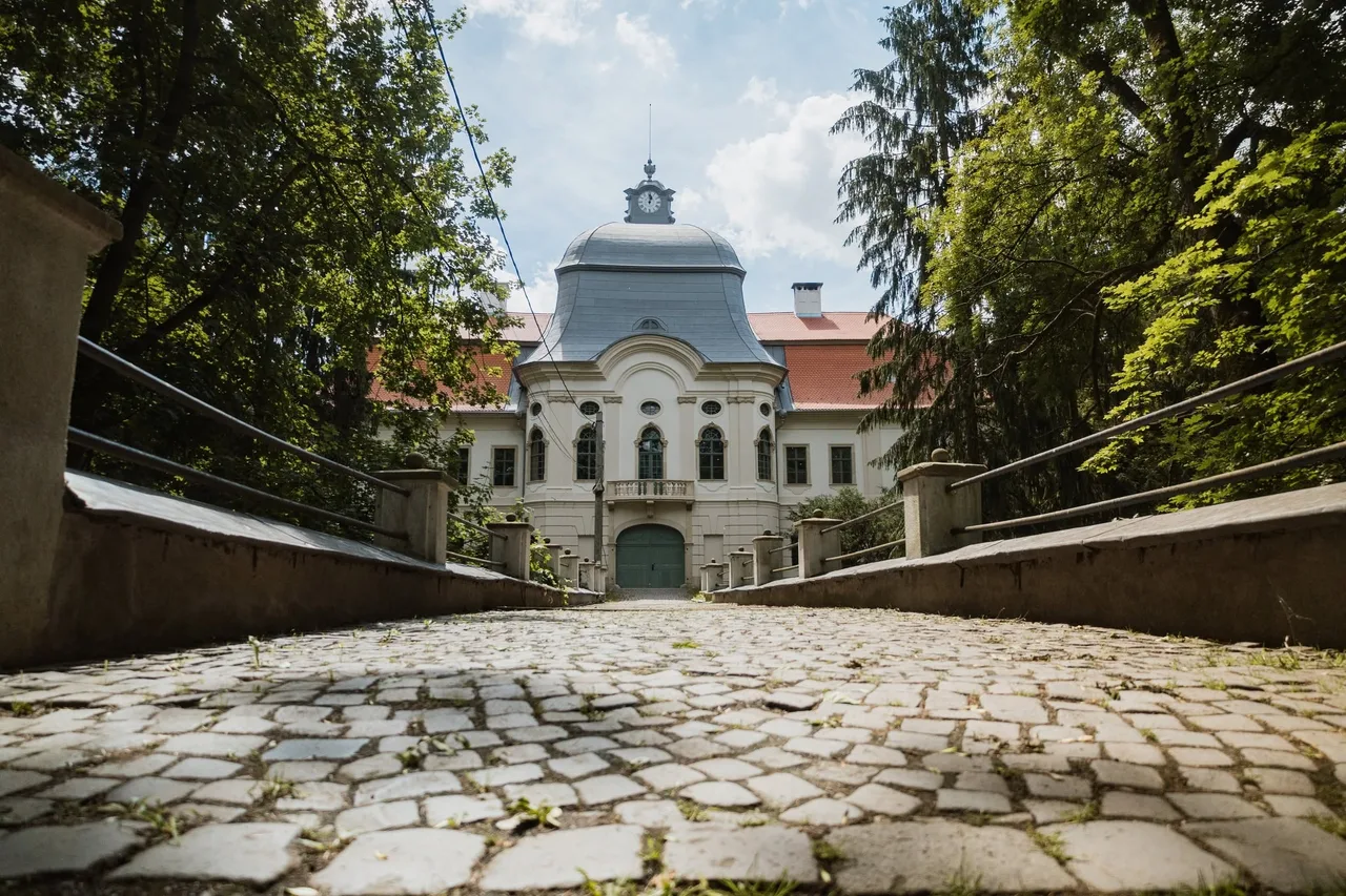 Kockakővel borított híd vezet a gernyeszegi Teleki-kastély főbejáratához, amelynek homlokzatán óra díszíti a kupolás tornyot, a bejárat mindkét oldalán fák magasodnak.