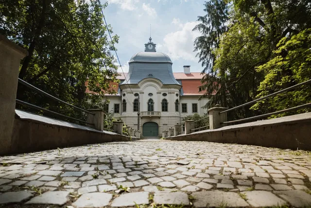 Kockakővel borított híd vezet a gernyeszegi Teleki-kastély főbejáratához, amelynek homlokzatán óra díszíti a kupolás tornyot, a bejárat mindkét oldalán fák magasodnak.