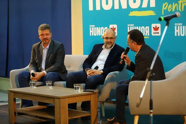 Három férfi ül fotelekben egy színpadon, UDMR/Kelemen Hunor kék hátterű logóval díszített teremben. Az egyikük mikrofont tart, és beszélget a másik kettővel, mintha interjút vagy panelbeszélgetést vezetne.