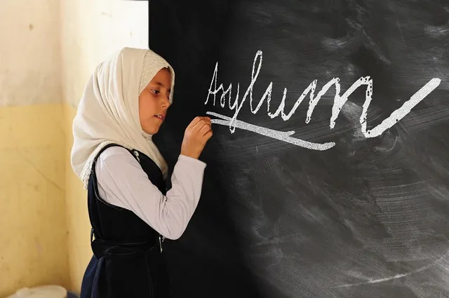 Fejkendőt viselő kislány iskolai egyenruhában krétával az „Asylum” szót írja egy fekete táblára.