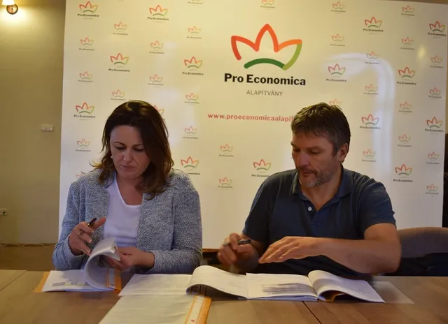 Egy nő és egy férfi ül egy asztalnál, dokumentumokat írnak alá tollal, mögöttük a Pro Economica Alapítvány logós molinó látható.