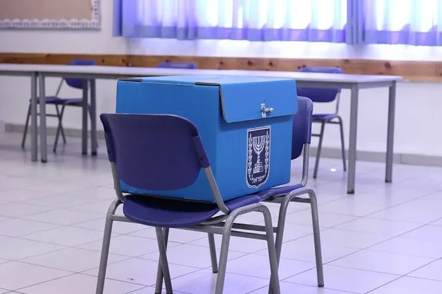Kék kartondoboz, rajta izraeli állami emblémával (menóra), egy szavazóurna, egy kék műanyag széken elhelyezve egy tanterem-szerű helyiségben, háttérben asztalok és székek.