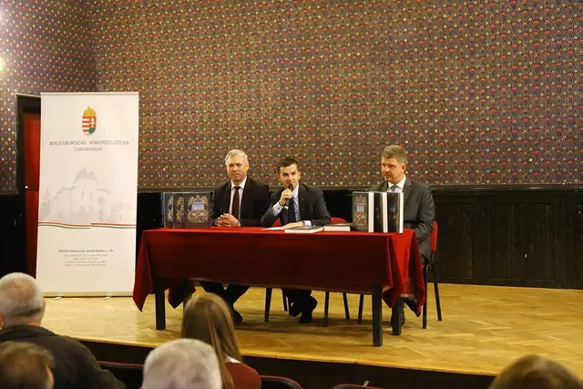 Három öltönyös férfi ül egy piros terítővel borított asztalnál egy díszteremben, középen mikrofonba beszél, mellettük könyvek láthatók az asztalon; a bal oldalon Magyarország Kormányzatának roll-up bannere áll, előtérben hallgatóság ül.