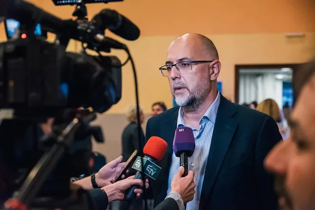 Kopasz, szemüveges, ősz szakállú férfi sötét zakóban nyilatkozik újságíróknak, akik mikrofonokat (köztük egy M1 feliratú mikrofont) tartanak felé, mellette videokamera rögzíti a jelenetet.