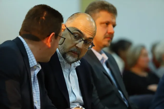 Öltönyös férfiak ülnek egymás mellett egy teremben, előtérben egy szemüveges, kopasz, ősz szakállú férfi hallgat figyelmesen, háttérben további emberek látszanak elmosódottan.