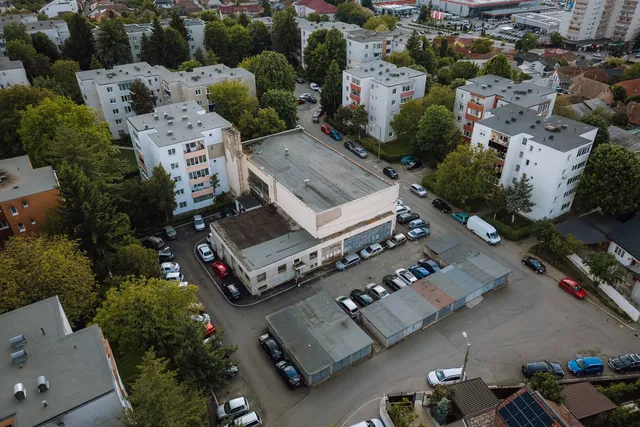 Légifotó egy lakóövezetről, ahol egy alacsony, lapos tetős épület körül parkoló autók és garázssorok láthatók, körülötte tömbházak és fák.