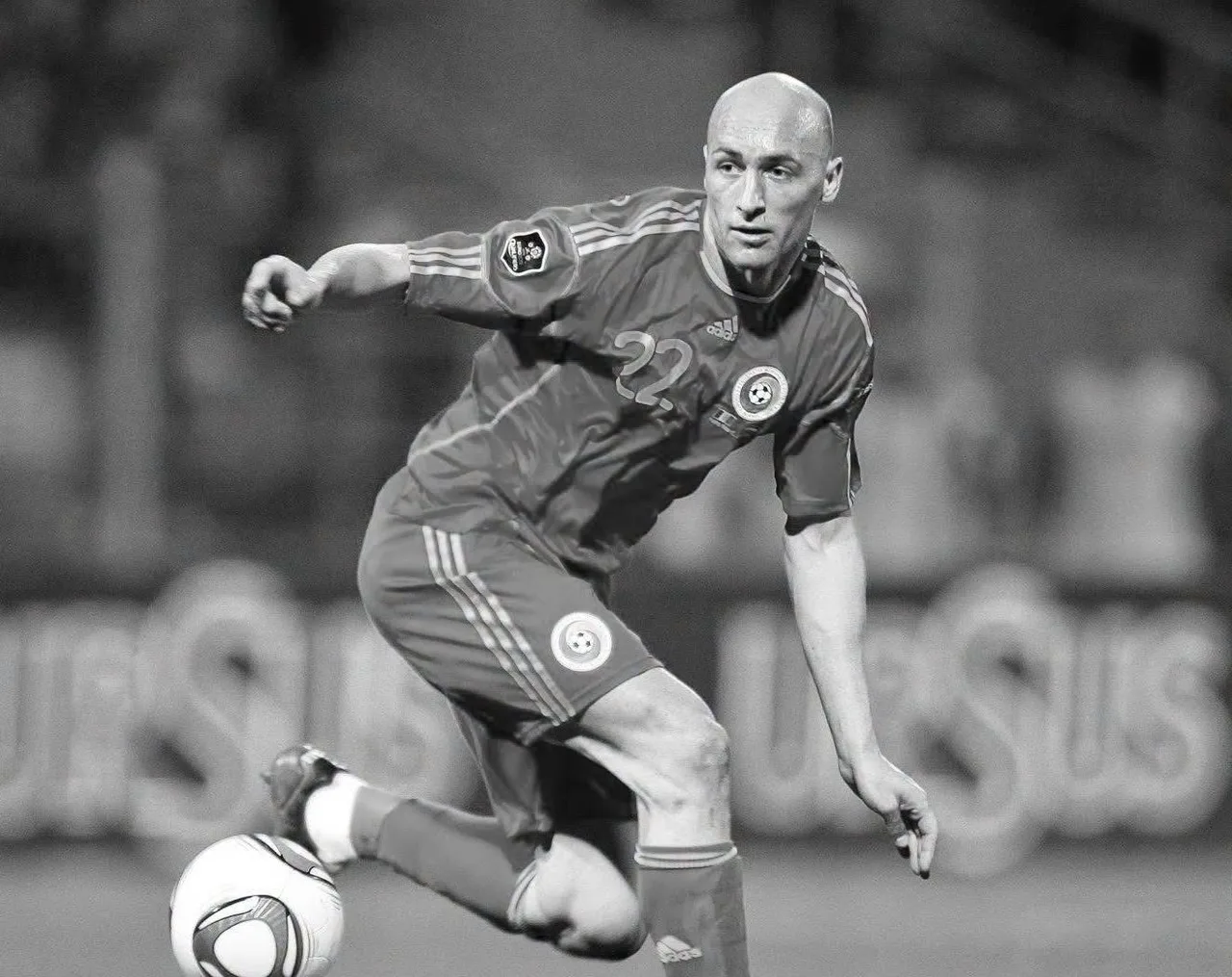 Fekete-fehér fotó egy futballistáról a román válogatott mezében, 22-es számmal, aki labdával a lábánál mozgás közben látható egy stadionban, háttérben elmosódott reklámtáblák.